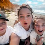 Three people on a sunny beach posing for a selfie, two kids and an adult, holding smooth rocks and smiling at the camera.