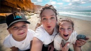 Three people on a sunny beach posing for a selfie, two kids and an adult, holding smooth rocks and smiling at the camera.
