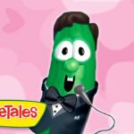 Green VeggieTales cucumber in a tuxedo, bow tie, and microphone, performing against a pink patterned background.