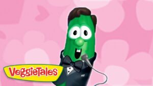 Green VeggieTales cucumber in a tuxedo, bow tie, and microphone, performing against a pink patterned background.
