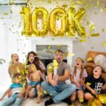 Family celebrating a milestone at home with large gold '100k' balloons and shower of confetti.