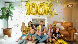 Family celebrating a milestone at home with large gold '100k' balloons and shower of confetti.