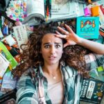Young woman lies on the floor surrounded by scattered books, notebooks, and school supplies, looking stressed. 125 characters max