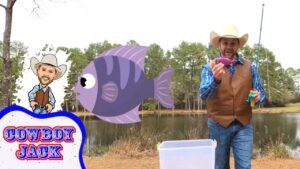 Cowboy wearing a hat and vest at a lake, holding a pink lure with a cartoon fish and ‘COWBOY JACK’ overlay.