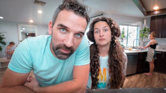 Couple leaning toward the camera in a bright modern kitchen, with a child in the background at a table and a woman at the sink nearby.