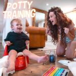 Toddler on a red potty chair smiles as mom cheers during potty training in a cozy living room, with toilet paper rolls and a color chart nearby.