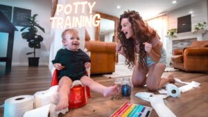 Toddler on a red potty chair smiles as mom cheers during potty training in a cozy living room, with toilet paper rolls and a color chart nearby.