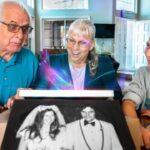 Three generations—an elderly man and woman with a young girl—smile as a wedding photo emerges from a device on the table amid a colorful glow, inside a cozy home.
