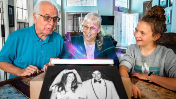 Three generations—an elderly man and woman with a young girl—smile as a wedding photo emerges from a device on the table amid a colorful glow, inside a cozy home.