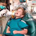Child patient reclined in a dental chair as a clinician in PPE works on their mouth with dental equipment nearby.