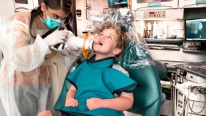 Child patient reclined in a dental chair as a clinician in PPE works on their mouth with dental equipment nearby.