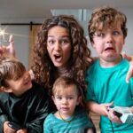 A mom and three young children making funny faces in a living room, pretending with toy hair clippers and scissors during playtime.