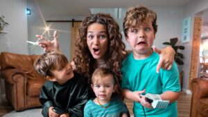 A mom and three young children making funny faces in a living room, pretending with toy hair clippers and scissors during playtime.