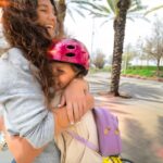 Two friends share a joyful hug outdoors; one wearing a pink bike helmet and a lavender backpack stands close as they smile at each other.