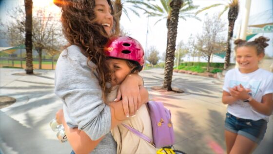 Two friends share a joyful hug outdoors; one wearing a pink bike helmet and a lavender backpack stands close as they smile at each other.