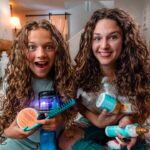 Two girls with long curly hair sit on the living room floor, smiling, holding assorted hair care products and brushes.