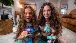 Two girls with long curly hair sit on the living room floor, smiling, holding assorted hair care products and brushes.