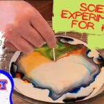 Hand swirls neon colors in a shallow dish for a kids' science experiment. A toothpick guides the mixing on a wooden table.