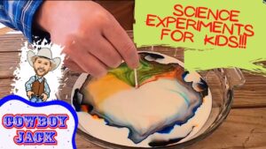 Hand swirls neon colors in a shallow dish for a kids' science experiment. A toothpick guides the mixing on a wooden table.