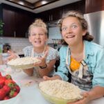 Two girls bake together in a bright kitchen, smiling while holding bowls of crumb-topped pie with fresh strawberries on the counter nearby.