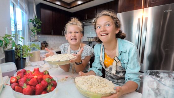 Two girls bake together in a bright kitchen, smiling while holding bowls of crumb-topped pie with fresh strawberries on the counter nearby.