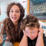 Woman with long curly hair speaks to a young boy who covers his ears in a bright classroom setting.