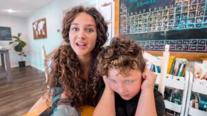 Woman with long curly hair speaks to a young boy who covers his ears in a bright classroom setting.