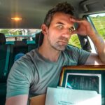 Man in a car resting his head on his hand, looking tired, with a framed document and papers in the front seat behind him.