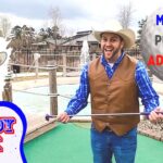 Man in a cowboy hat and brown vest holds a putter on a mini-golf course, with a cartoon cowboy cutout and 'Cowboy Jack' sign in the scene.