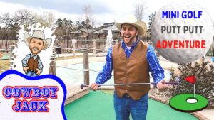 Man in a cowboy hat and brown vest holds a putter on a mini-golf course, with a cartoon cowboy cutout and 'Cowboy Jack' sign in the scene.
