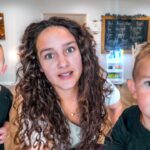 Mother with curly hair leaning toward the camera, flanked by two young boys in green shirts in a bright playroom; world map and chalkboard in the background.