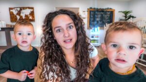 Mother with curly hair leaning toward the camera, flanked by two young boys in green shirts in a bright playroom; world map and chalkboard in the background.