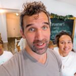 Man in the foreground takes a selfie with two smiling people in a bright, cozy living space behind him