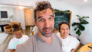 Man in the foreground takes a selfie with two smiling people in a bright, cozy living space behind him