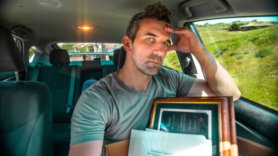 Man in a car, resting his forehead on his hand while holding a framed certificate and papers on his lap.