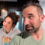 Man in a green shirt and a woman in a hoodie lean close together in a modern kitchen, smiling at the camera (selfie).