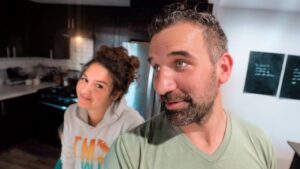 Man in a green shirt and a woman in a hoodie lean close together in a modern kitchen, smiling at the camera (selfie).