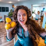 Woman with long brown hair holding three lemons toward the camera, smiling in a bright kitchen with kids playing in the background.