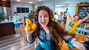 Woman with long brown hair holding three lemons toward the camera, smiling in a bright kitchen with kids playing in the background.
