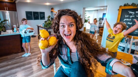 Woman with long brown hair holding three lemons toward the camera, smiling in a bright kitchen with kids playing in the background.