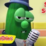 Larry the Cucumber from VeggieTales wearing a hat and suit, singing into a vintage microphone with 'Silly Songs' title in the corner.