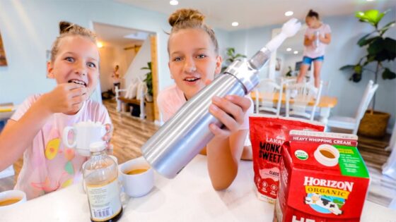Two young girls sit at a bright cafe table; one proudly holds a large metal shaker as they prepare drinks, with milk and cups on the table.