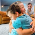 Two young children share a warm hug in a bright living room, with a man seated in the background.