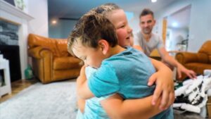 Two young children share a warm hug in a bright living room, with a man seated in the background.