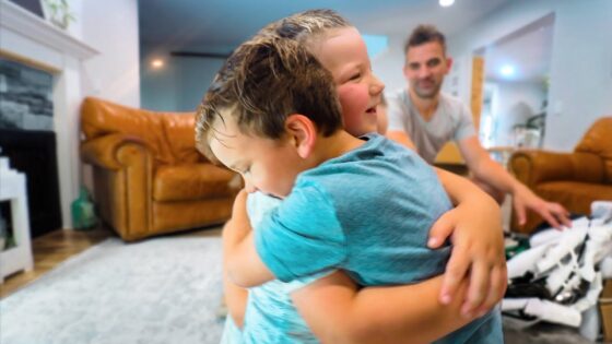 Two young children share a warm hug in a bright living room, with a man seated in the background.