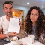 Man and woman at a cafe table with ice cream; the man holds a document and pen toward the camera.