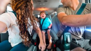 People riding in a minivan; a child in the middle seat is crying while two adults in the front seats drive or interact nearby.