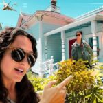 Woman wearing sunglasses takes a selfie in front of a bright turquoise house, while a man in a green flight suit stands on the porch in the background under a sunny sky.