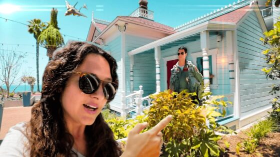 Woman wearing sunglasses takes a selfie in front of a bright turquoise house, while a man in a green flight suit stands on the porch in the background under a sunny sky.