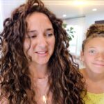 Mother with long curly hair and daughter with a bun smiling in a bright living room, indoors in a home setting.